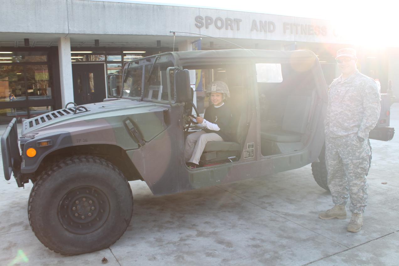 A boy sits in a jeep with a soldier nearby.